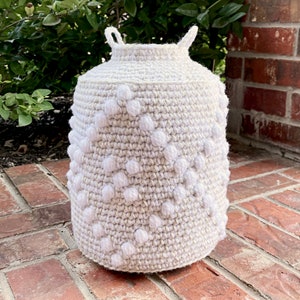 May include: A white crocheted basket with a textured pattern and white pom-poms. The basket is sitting on a brick surface.