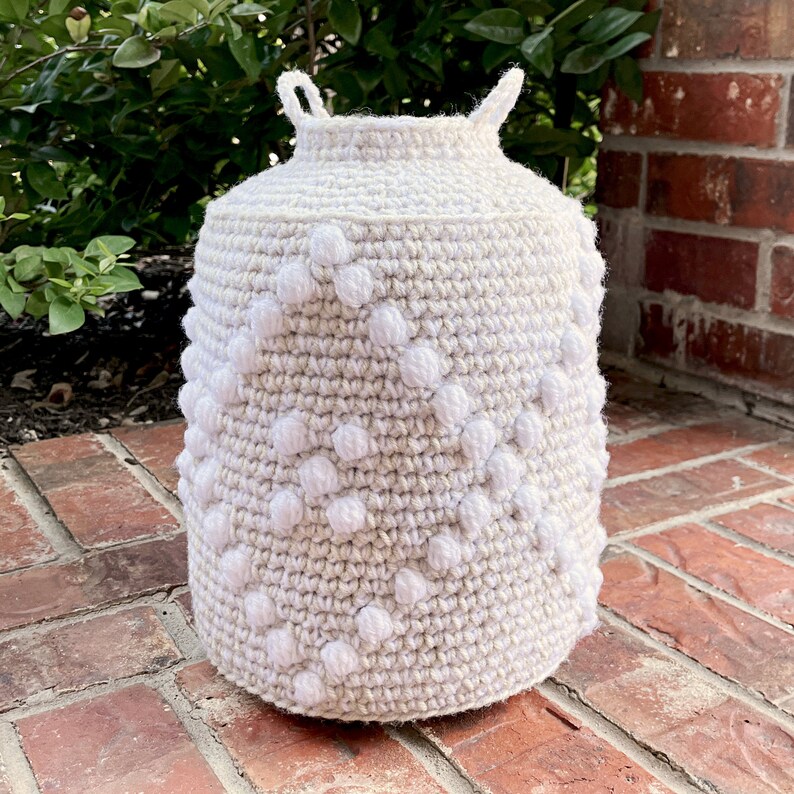 May include: A white crocheted basket with a textured pattern and white pom-poms. The basket is sitting on a brick surface.