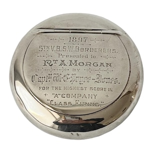 Puede incluir: Una medalla redonda de plata con una inscripción que dice: "1897 5TH V.B.S.W.BORDERERS. Presented to R.T.A. MORGAN by Capt. W.G. Pryce-Jones. FOR THE HIGHEST SCORE IN "A" COMPANY CLASS FIRING"