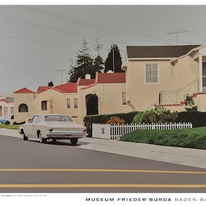 May include: A vintage white car drives down a street lined with beige houses with red tile roofs. A white picket fence and green bushes border the sidewalk. The sky is overcast. The artwork is displayed at the Museum Frieder Burda Baden-Baden.