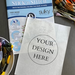 May include: A white sheet of printable fabric with the words "YOUR DESIGN HERE" printed in a circle in the center. The fabric is designed for use with embroidery, cross stitch, punch needle, and quilting.