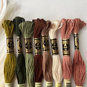 May include: A collection of DMC Mouline Special 25 embroidery floss in various colors, including olive green, brown, and peach. Each skein is labeled with a product code and contains 8 meters (8.7 yards) of thread, perfect for cross-stitch and embroidery projects.