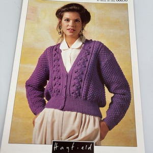May include: A knitting pattern leaflet titled DOUBLE KNIT 00630 by Hayfield, featuring a purple V-neck cardigan with a textured bobble stitch pattern, ribbed cuffs, and a button-front closure, designed for chest sizes 76-102cm.