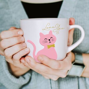 May include: A white ceramic mug with a pink cat illustration, a gold bow tie, and the words "Love You" in gold script. The mug has a curved handle. The mug is held by a person wearing a grey jumper.