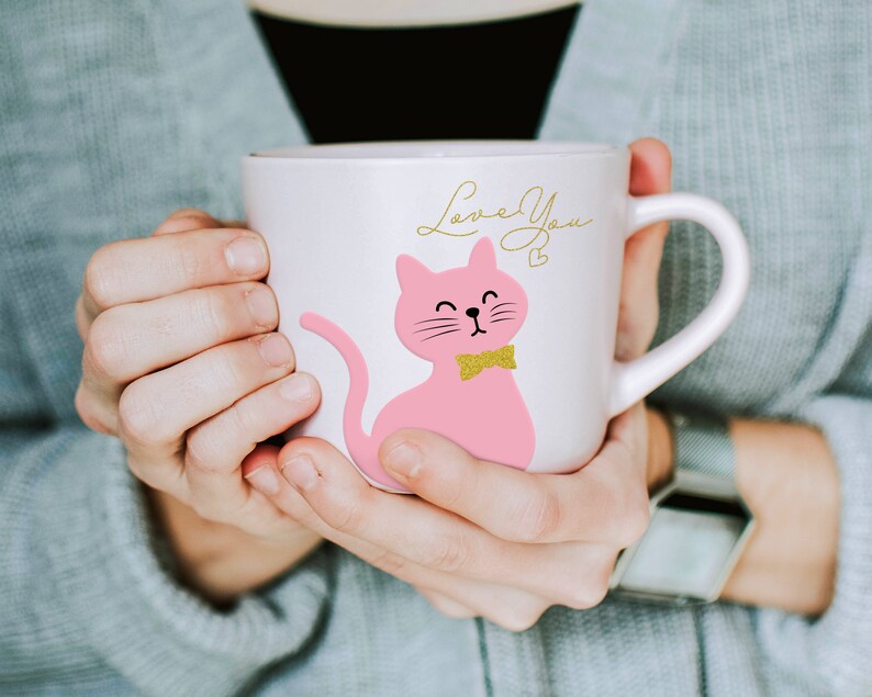 May include: A white ceramic mug with a pink cat illustration, a gold bow tie, and the words "Love You" in gold script. The mug has a curved handle. The mug is held by a person wearing a grey jumper.