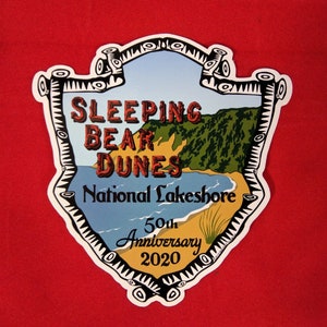 May include: A white sticker with a black outline in the shape of a shield. The sticker features a colorful illustration of Sleeping Bear Dunes National Lakeshore with the text "Sleeping Bear Dunes National Lakeshore 50th Anniversary 2020".