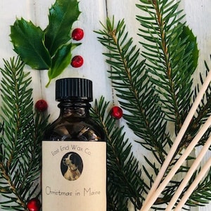 May include: A brown glass bottle with a black lid and a white label that reads "East End Wax Co. Christmas in Maine Reed Diffuser hand poured on the coast of Maine". The bottle is surrounded by green pine branches and red berries.