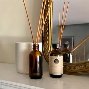 May include: A brown glass bottle with a brown label that reads "Leaf Pepper" and "Reed Diffuser" with a small illustration of a dog. The bottle is filled with a clear liquid and has a set of brown wooden sticks in it. The bottle is sitting on a white surface next to a white ceramic cylinder.
