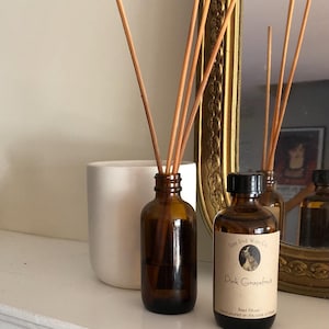 May include: A brown glass bottle with a white label that reads "East End Wax Co. Pink Grapefruit Reed Diffuser Hand poured on the coast of Maine". The bottle is filled with a clear liquid and has wooden sticks inserted into it. The bottle is sitting on a white surface next to a white ceramic container.