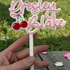 May include: A layered cake topper with the names "Presley Blake" in pink script, outlined in white. Two red cherries with green stems are on the left. The topper is held by a white stick, ideal for a birthday or celebration.