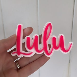 May include: A pink acrylic name sign with the word "Lulu" in a cursive font.
