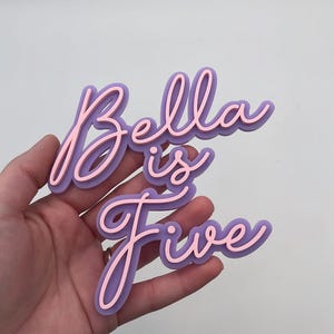 Peut inclure: Une enseigne superposée affichant le texte Bella is Five en écriture cursive. La couche supérieure est d'une couleur rose clair, tandis que la couche inférieure est violette unie, créant un effet dimensionnel avec le lettrage empilé.