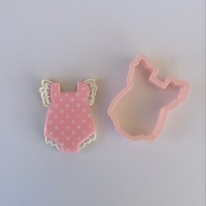 Peut inclure: Emporte-pi&egrave;ce rose en forme de body b&eacute;b&eacute; avec des pois blancs et une bordure en dentelle blanche. L'emporte-pi&egrave;ce est sur une surface blanche.
