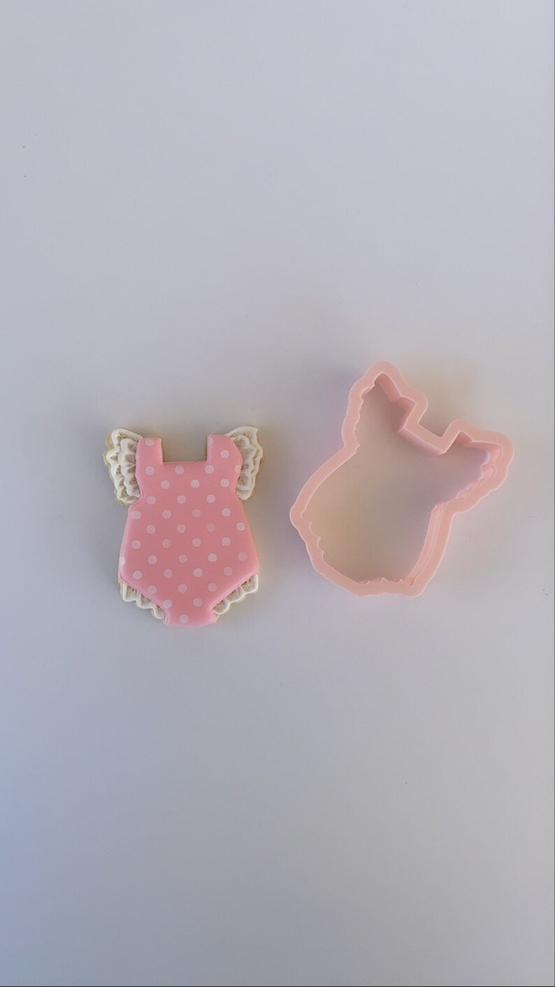 Peut inclure: Emporte-pi&egrave;ce rose en forme de body b&eacute;b&eacute; avec des pois blancs et une bordure en dentelle blanche. L'emporte-pi&egrave;ce est sur une surface blanche.