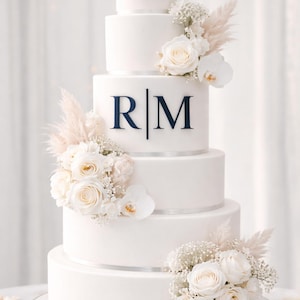 Puede incluir: Una tarta de boda blanca de cinco pisos decorada con rosas blancas, orquídeas y hierba de la Pampa. La tarta presenta las iniciales "RM" en azul marino en el tercer piso y está adornada con cinta plateada.