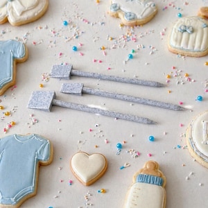 May include: An assortment of baby-themed sugar cookies, including a onesie, baby bottle, and heart-shaped cookie, decorated with blue and white icing. Three gray knitting needles are also present, along with colorful sprinkles.