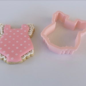 Peut inclure: Un biscuit rose et blanc d&eacute;cor&eacute; d'un motif de barboteuse de b&eacute;b&eacute; &agrave; pois. Le biscuit est &agrave; c&ocirc;t&eacute; d'un emporte-pi&egrave;ce en plastique en forme de barboteuse de b&eacute;b&eacute;.