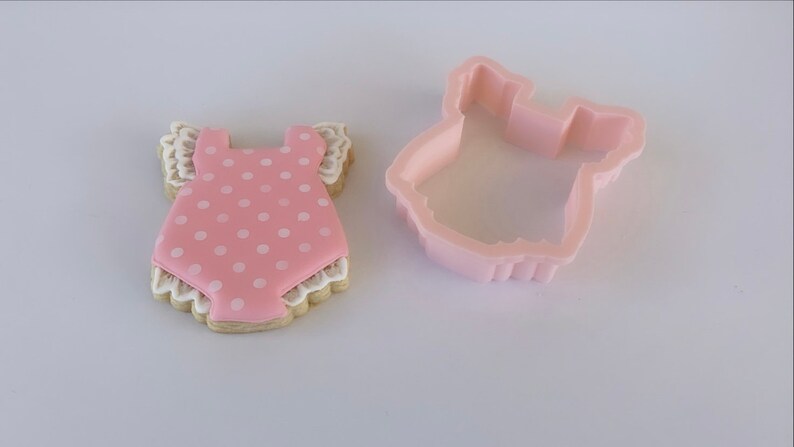 Peut inclure: Un biscuit rose et blanc d&eacute;cor&eacute; d'un motif de barboteuse de b&eacute;b&eacute; &agrave; pois. Le biscuit est &agrave; c&ocirc;t&eacute; d'un emporte-pi&egrave;ce en plastique en forme de barboteuse de b&eacute;b&eacute;.