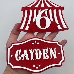 May include: A red and white circus tent topper featuring the number 6 and a separate red and white plaque with the name CAYDEN in white lettering. Both items are constructed with layered materials and feature a bold, high-contrast colour scheme.