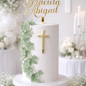 May include: A cylindrical white cake decorated with green ivy leaves and small white pearls, featuring a gold cross on the front and a gold acrylic cake topper with the names Graciela Abigail in a cursive script font, set on a white stand.