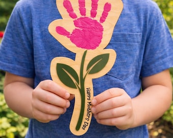 May include: A wooden flower-shaped craft with a pink handprint in the center. The flower has a stem and leaves, with the text "Happy Mother's Day" printed on the stem. The craft is held by a person wearing a blue shirt.
