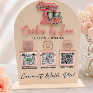 Pode incluir: Uma placa bege em arco com um gráfico de batedeira rosa com flores, o texto Cookies by Sam CUSTOM COOKIES e três ícones em ouro rosa com códigos QR para pagamentos e redes sociais, terminando com o texto Connect With Us!