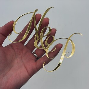 Puede incluir: Un recorte de acrílico de color dorado que presenta las letras N y P conectadas por un ampersand en una fuente cursiva fluida. La superficie de las letras es reflectante y lisa, creando una apariencia metálica con bordes definidos.