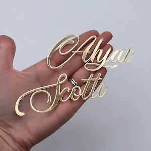 Puede incluir: Dos nombres de acrílico dorado, "Ahyan" y "Scotti", se muestran en una fuente cursiva. Los nombres se sostienen en la mano de una persona.