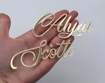 Può includere: Due ritagli di nomi in acrilico dorato e riflettente che riportano i nomi Ahyai e Scotti in un carattere corsivo. Le lettere sono collegate in uno stile fluido e decorativo con una finitura superficiale liscia e lucida e bordi netti.