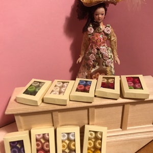 Peut inclure: Une poupée en costume d'époque et chapeau se tient derrière plusieurs petites boîtes. Chaque boîte contient une collection de petites bobines de fil colorées, parfaites pour les travaux manuels de maison de poupée ou les projets miniatures. Les boîtes sont disposées sur une surface en bois.