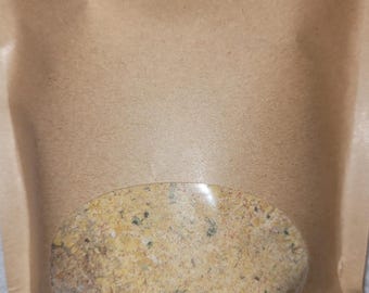 May include: A light brown kraft paper stand-up pouch featuring a clear oval window in the center. The pouch is filled with a coarse, dry, golden-yellow seasoning blend containing visible flecks of green herbs and small dark particles throughout.