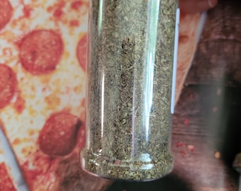 May include: A clear glass jar filled with dried green herbs. The jar has a red lid and is sitting on a pizza box.