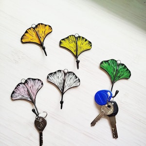 Könnte beinhalten: Fünf Ginkgo-Blatt-Schlüsselanhänger in Gelb, Rosa, Grün und Schwarz-Weiß. Jeder Schlüsselanhänger hat eine kleine Schlaufe für Schlüssel und einen schwarzen Stiel. Ein Schlüsselanhänger hält einen Satz Schlüssel.