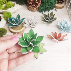 May include: Assortment of six stained glass succulent decorations in various colors, including green, white, blue, orange, and a darker green. The succulents are arranged on a white surface.