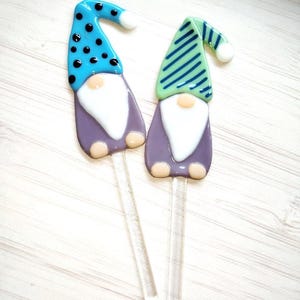 May include: Two glass garden gnome plant stakes. One gnome has a blue hat with black polka dots, the other has a green hat with blue stripes. Both have white beards, purple bodies, and clear stems.