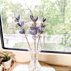 May include: A clear glass vase holds three stems of handcrafted, stained glass flowers. The flowers have blue petals and metallic stems. The vase sits on a white surface in front of a window with a blurred green background.