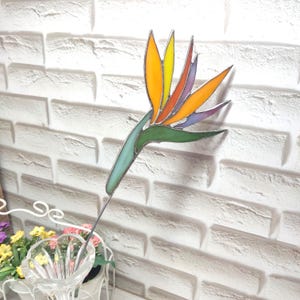 Peut inclure: Une fleur de paradisier en vitrail aux couleurs vives. La fleur présente des pétales jaunes, oranges et violets, avec une tige verte et une tige métallique pour l'affichage. L'œuvre est placée devant un mur de briques blanches.