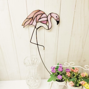 Op de afbeelding: Een glas-in-lood flamingo ornament met lichtroze en perzikkleurige glasstukken die bij elkaar worden gehouden door donkere metalen soldeerlijnen. De flamingo staat in een staande houding met een lange nek en een gebogen snavel op een dunne metalen staaf.