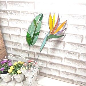 Può includere: Un fiore uccello del paradiso in vetro colorato e una foglia verde separata, entrambi montati su sottili steli di metallo. Il fiore presenta vivaci petali in vetro giallo, arancione e viola con una base verde, mentre la foglia è composta da varie tonalità di vetro verde.