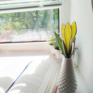 May include: A white, textured vase holding a yellow and green stained glass flower. An open book with a blue bookmark rests on a white surface, near a window with blinds. Small potted plants are visible in the background.