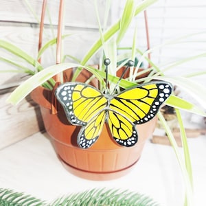 Può includere: Un ornamento a forma di farfalla in vetro colorato con ali giallo brillante, venature nere e bordi neri con puntini bianchi. La farfalla ha due sottili antenne in metallo ed è fissata a un vaso pensile in plastica marrone con una pianta verde.