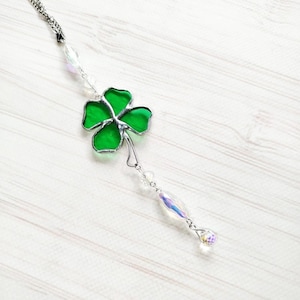 May include: A green stained glass four-leaf clover suncatcher with a silver-colored metal frame and chain. The clover is accented with clear iridescent beads and a teardrop-shaped crystal. The suncatcher hangs on a white wooden surface.