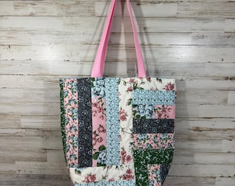 May include: A quilted tote bag with a patchwork design. The bag features a variety of floral patterns in pink, blue, and green, with a solid pink strap. The bag is hanging against a light wood-paneled wall.