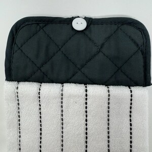 May include: A black quilted kitchen towel holder with a white button closure. The towel is white with vertical black dotted stripes. This kitchen accessory is designed for hanging towels.