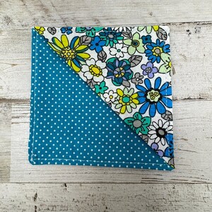 May include: Square fabric coaster with a teal polka dot pattern and a floral print. The floral print features blue, yellow, and white flowers on a white background. The coaster is edged with white stitching.