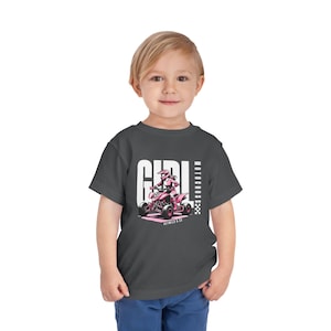 Puede incluir: Una camiseta gris oscuro con un gráfico rosa y blanco de una niña conduciendo un ATV. El texto "GIRL" y "MOTOCROSS" está impreso verticalmente en la camiseta. El texto "REWILD & CO" está impreso debajo del gráfico.