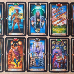 May include: A collection of tarot cards with black borders and gold accents. Each card features detailed illustrations and titles such as "Ace of Pentacles," "The Magician," and "The Sun." The cards are arranged in rows, showcasing various scenes and figures.