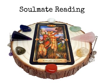 Op de afbeelding: Een tarotkaartopstelling met de kaart Ridder van Bekers, omringd door kristallen en een pendel op een houten schijf. De tekst "Soulmate Reading" staat boven de kaart. De kaart toont een ridder te paard.