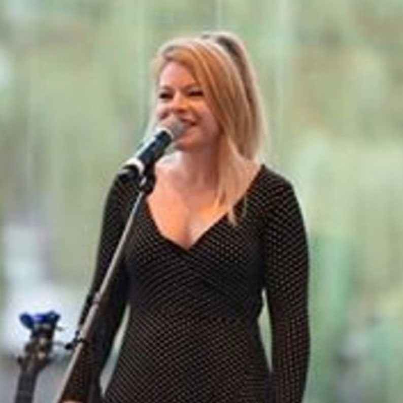 May include: A woman with blonde hair sings into a microphone. She wears a black dress with white polka dots and long sleeves. The microphone is attached to a stand, and a guitar is visible in the background.