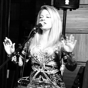 May include: A black and white image of a person singing into a microphone. The person is wearing a patterned dress with long sleeves and has long blonde hair. The person is gesturing with their hands.